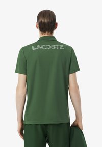 Polo verde de manga corta, con un logotipo blanco de "LACOSTE" en la espalda. Confeccionado con tela suave y cuello clásico.