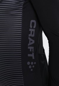 Černý sportovní top s vertikálním pruhem na jedné straně, s velkým logem "CRAFT" a kruhovým designovým akcentem ve tmavší barvě.