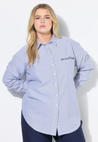 Camicia a righe blu e bianche con orlo curvo, dotata di maniche lunghe e testo ricamato vicino al petto. Tessuto morbido.