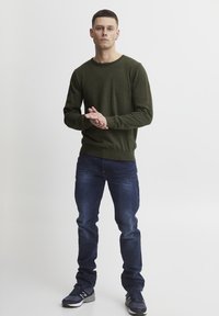 Maslinasto zeleni pleteni džemper s okruglim izrezom, uparen s tamnoplavim trapericama. Model nosi sive tenisice. Jednostavan dizajn, bez uzoraka ili ukrasa.