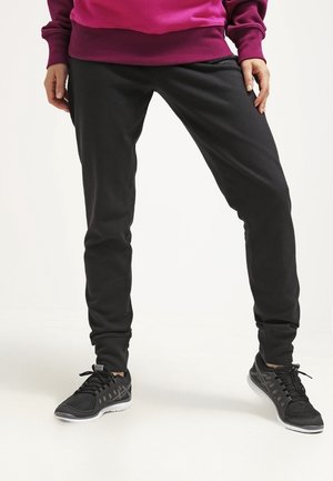 Person trägt schwarze Jogginghosen, schwarze Sneaker mit weißen Sohlen und einen lila Pullover, steht mit gespreizten Beinen vor weißem Hintergrund.