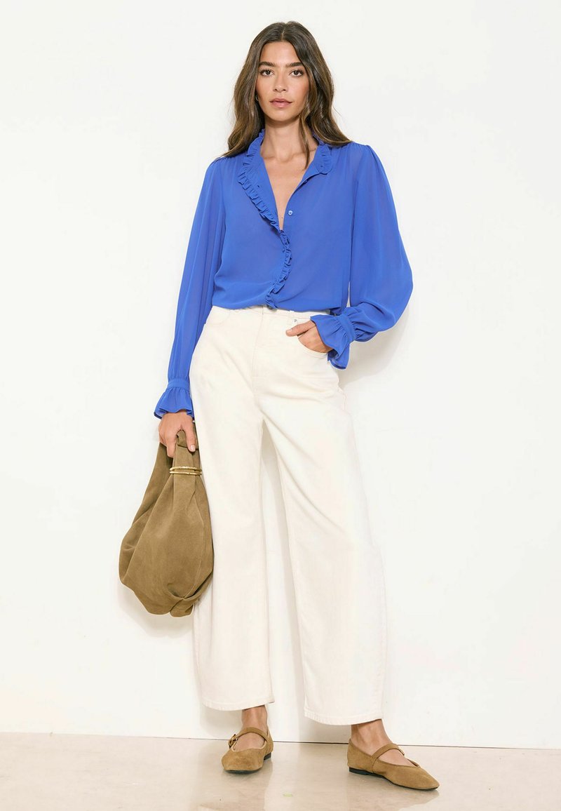 Sieviete valkā zilu bārkstainu blūzi, baltas platas bikses, brūnas dzīvokļveida kurpes un tur lielu brūnu zamšādas somu, stāvot pret balto fonu.