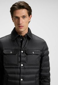 Svart vadderad jacka med krage, två bröstfickor och knappstängning. Slät textur med glänsande finish och kontrasterande silverfärgad besättning.