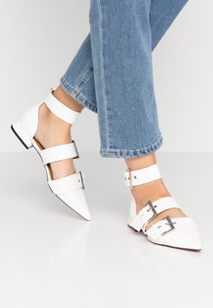 Białe baleriny z noskiem w szpic, podwójnymi paskami wokół kostki i metalowymi klamrami. Gładka faktura i niski, blokowy obcas. Noszone z niebieskimi, szerokimi spodniami typu jeans.