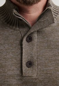 Primo piano di un uomo che indossa un maglione lavorato a maglia marrone con un alto colletto, due bottoni scuri e una cerniera parzialmente aperta.