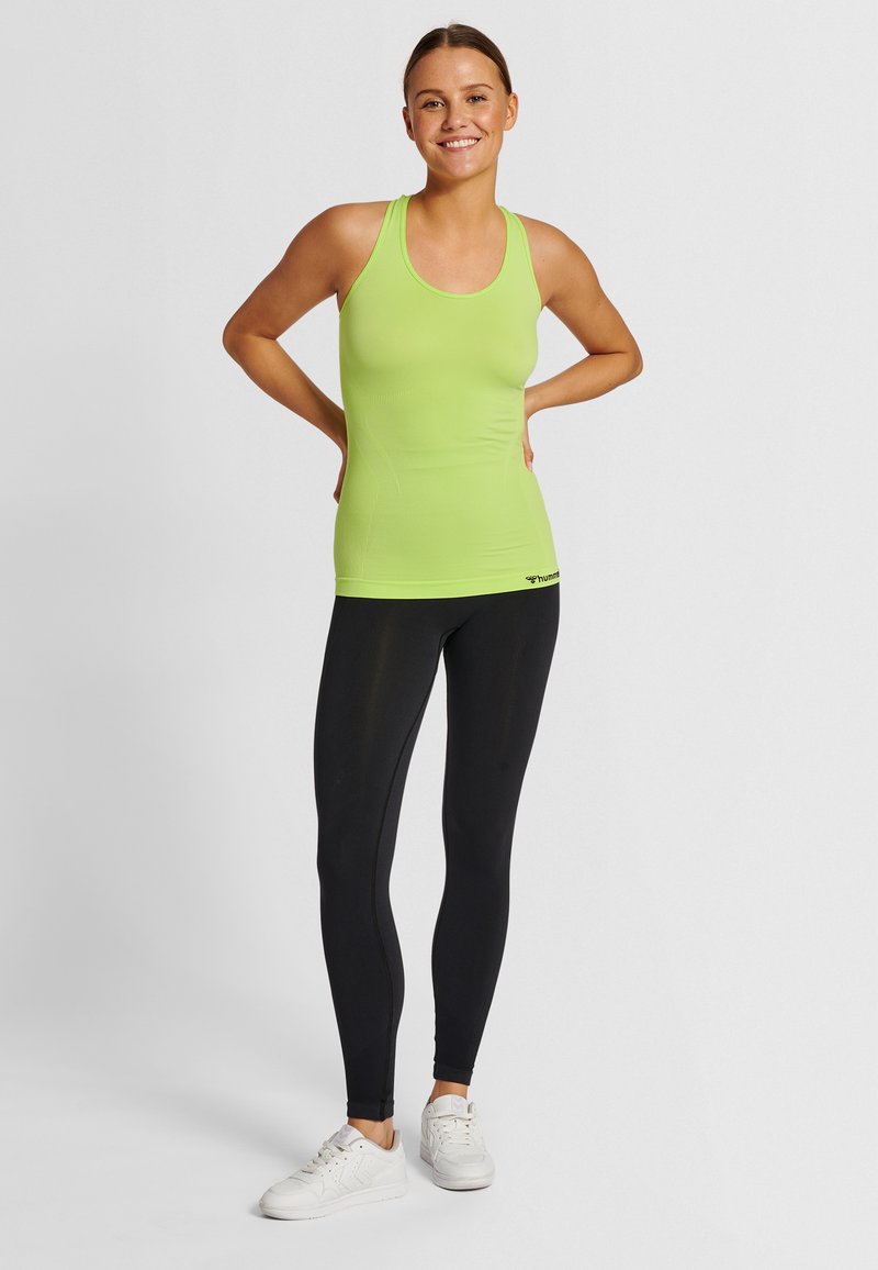 Smiling woman wearing a lime green sleeveless athletic top, black leggings, and white sneakers, standing with hands on hips on a neutral background.