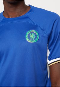 Camisa de fútbol azul con tela texturada, que presenta un logo circular del club en verde con un león y el texto "Chelsea Football Club". Acentos en blanco y dorado.