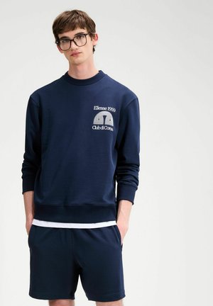 Młody mężczyzna w okularach, ubrany w granatową bluzę i pasujące szorty, pod którymi widać białą koszulkę, stoi z rękami w kieszeniach.