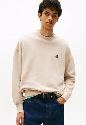 Sudadera beig de cuello redondo con puños de canalé, que presenta un pequeño parche de logo multicolor en el pecho, combinada con unos jeans de mezclilla azul y un cinturón negro.
