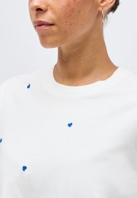 Γυναίκα με φακίδες, που φοράει λευκό πουκάμισο με μικρές μπλε κεντημένες καρδιές και ασημένιο κρίκο σκουλαρίκι σε απλό φόντο.