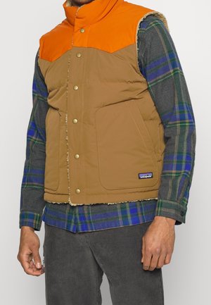 Mann trägt eine beige-orangefarbene, ärmellose, gepolsterte Weste über einem grün-blau karierten Hemd und graue Cordhosen.