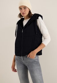 Marinblå pufferväst med huva av fuskpäls, dragkedja fram och två sidofickor. Bärs över en lätt tröja och kombineras med grå jeans.