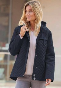 Schwarze gesteppte Jacke mit Kapuze, ausgestattet mit Druckknöpfen und seitlichen Taschen. Über einem leichten Pullover getragen, mit einer strukturierten Oberfläche.