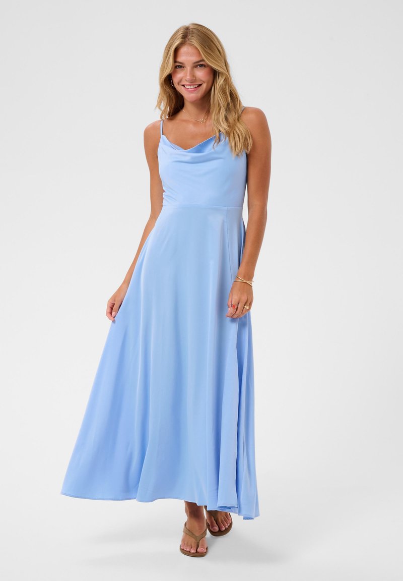 Femme souriante aux longs cheveux blonds portant une robe longue sans manches bleu clair et des sandales, debout devant un fond uni.