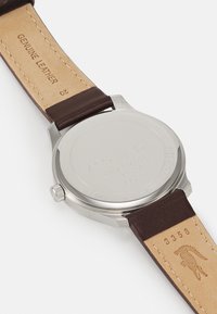 Bracelet de montre en cuir marron avec des coutures blanches, dos en acier inoxydable gravé avec la marque et les détails, texture lisse, design minimaliste.