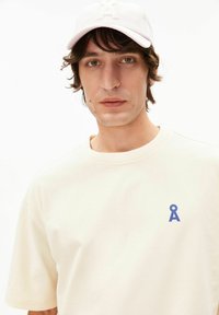 Beige T-shirt met korte mouwen en een blauw logo op de linkerborst, gecombineerd met een lichtroze baseballpet met reliëfletters.
