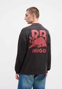 Maglietta nera a maniche lunghe con una grande grafica di un toro rosso e il testo "PRHUGO" sul retro. Vestibilità morbida con polsini a coste e texture soffice.