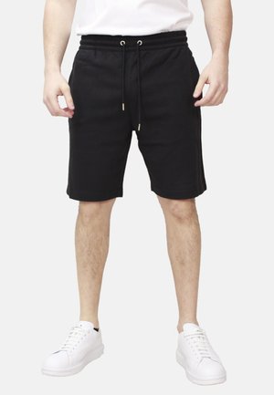 Uomo che indossa pantaloncini neri con coulisse e sneakers bianche, in piedi su uno sfondo semplice con le mani rilassate ai lati.