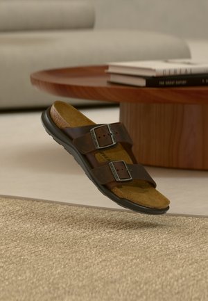 Hnedá kožená sandála s dvoma nastaviteľnými prackami vznášajúca sa nad tkaným béžovým kobercom v obývačke.