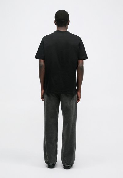 T-shirt de algodão preto com decote redondo e mangas curtas, combinado com jeans de perna larga cinza escuro, exibido pela parte de trás sobre um fundo plain.