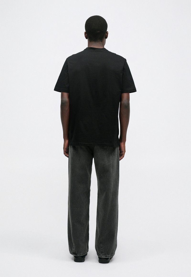 T-shirt en coton noir avec un col rond et des manches courtes, associé à un pantalon en jean large gris foncé, affiché de dos sur un fond uni.