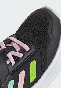 Chaussure de sport noire avec tige en mesh, lacets roses et trois accents colorés (vert, rose et teal) sur le côté. Surfaces texturées visibles.