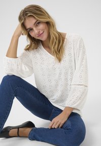 Top blanc avec texture à œillets et encolure en V, doté de manches ballon. Associé à un jean en denim bleu et des chaussures à talons noires.