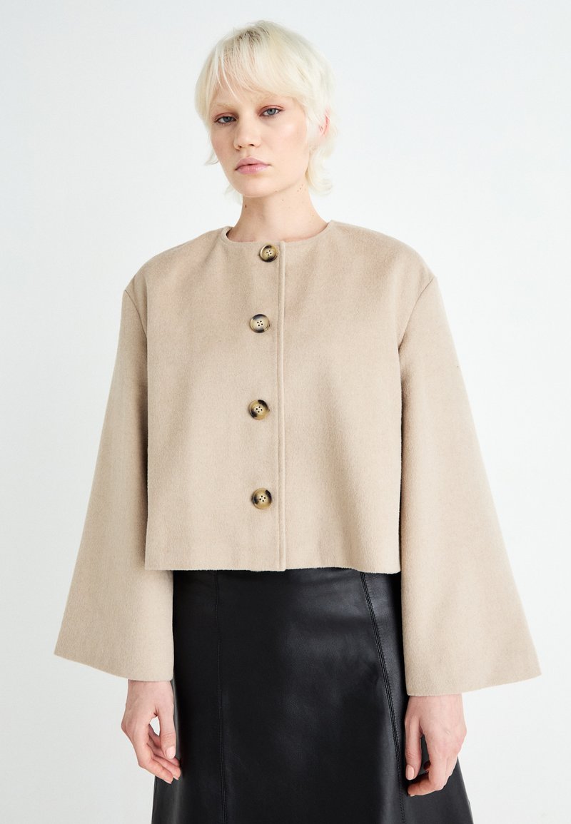 Giacca cropped beige realizzata in morbido tessuto, con colletto arrotondato e cinque grandi bottoni dorati lungo il frontale. Maniche lunghe svasate.