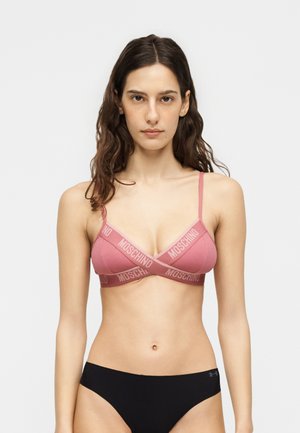 Kobieta z brązowymi falującymi włosami sięgającymi talii, ubrana w różowy bralet Moschino z logo na ramiączkach i czarną bieliznę, stojąca na jednolitym tle.