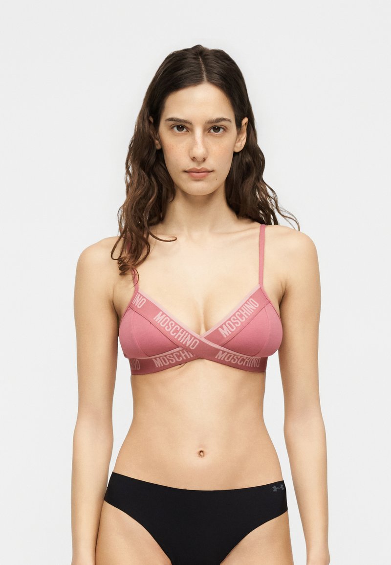 Γυναίκα με καστανά κυματιστά μαλλιά μέχρι τη μέση που φορά ροζ bralette Moschino με ιμάντες λογότυπου και μαύρο εσώρουχο, στέκεται μπροστά από απλό φόντο.