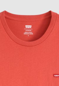 T-shirt en coton rouge avec un col rond, arborant un petit écusson blanc avec le logo Levi's sur le côté gauche. Étiquette visible à l'intérieur du col. Texture simple.