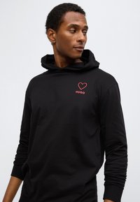 Μαύρο φούτερ με κουκούλα που διαθέτει κόκκινη καρδιά και λογότυπο "HUGO" στο στήθος, κατασκευασμένο από μαλακό, λείο ύφασμα με μακριά μανίκια.