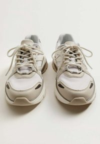 Chaussures de sport beige et blanches avec tissu en mesh, superpositions texturées, forme arrondie et design à lacets avec accents minimaux.