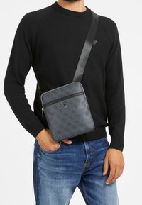 Ein Mann trägt einen schwarzen Pulli und blaue Jeans. Er hält eine graue Umhängetasche mit einem gemusterten Design und einem dreieckigen Logo auf der Vorderseite.