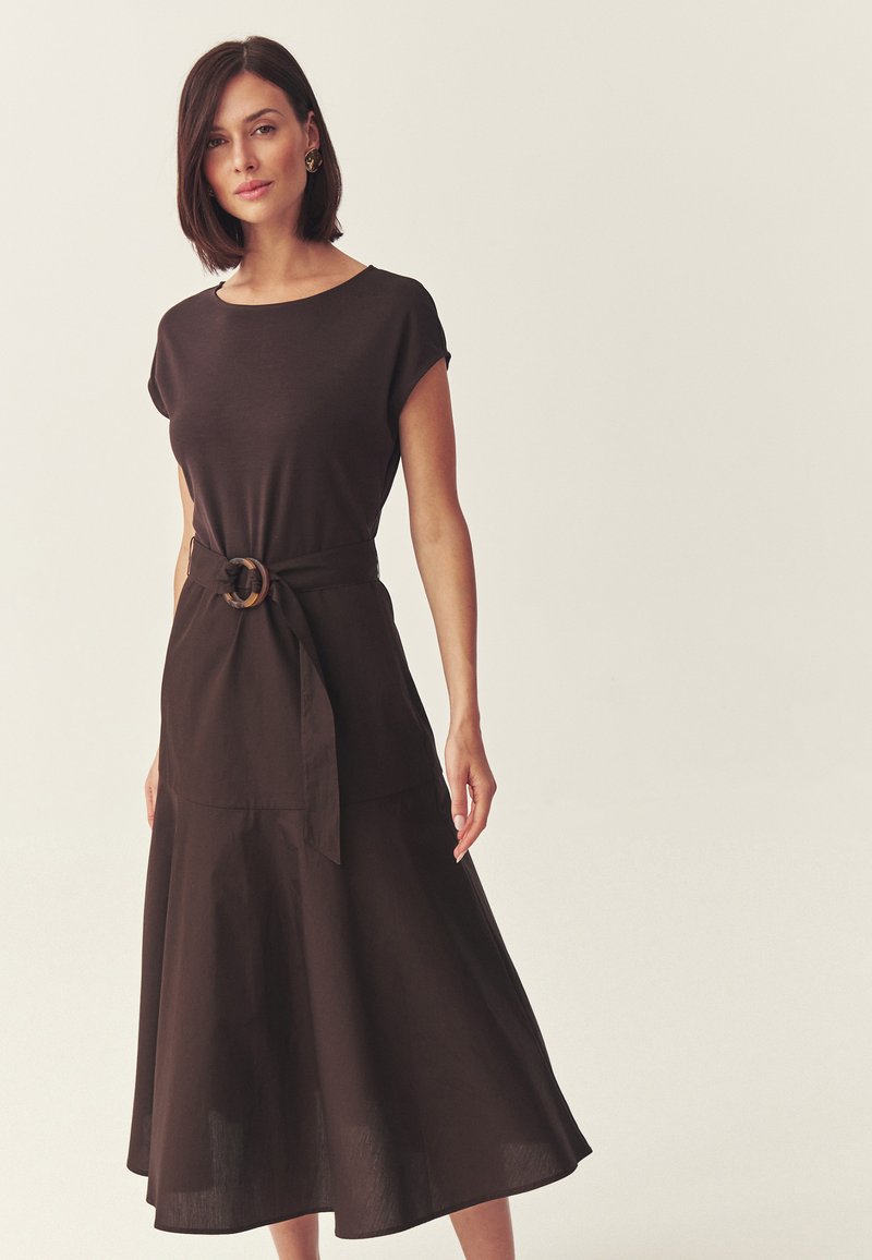 Femme aux cheveux courts et foncés portant une robe midi marron foncé ceinturée, à manches courtes, debout devant un fond clair uni.