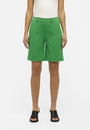 Femme portant un short vert vif sur mesure arrivant aux genoux, un haut blanc sans manches et des chaussures noires ouvertes aux orteils, debout devant un fond blanc.