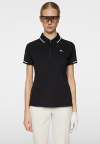 Svart pikétröja med dragkedja i halsen, vita detaljer på ärmarna, och en logotyp på bröstet. Bärs tillsammans med ljusa byxor och en golfhandske.