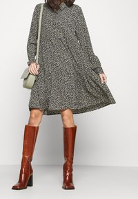 Abito nero a fiori con colletto e maniche lunghe, caratterizzato da una gonna fluida. Abbinato a stivali alti marroni e una borsa a tracolla verde.