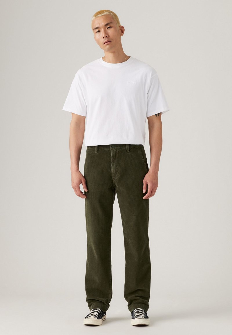 White short-sleeved t-shirt worn with dark green corduroy pants. Pants have a straight fit, visible stitching, and side pockets. Black and white sneakers.