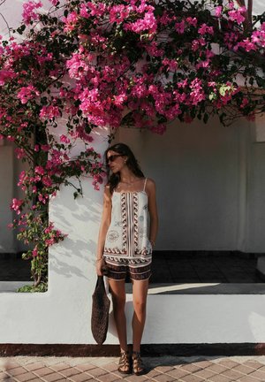 Žena v vzorovaných šatách a sandáloch stojí pod žiarivou ružovou bougainvilleou na bielom múre a drží tmavú pletenú tašku.