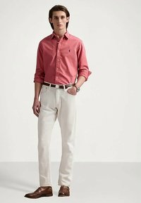 En ung man i en korallfärgad skjorta med knappar, vita byxor med ett brunt bälte, bruna loafers och en armbandsklocka, står mot en enkel bakgrund.