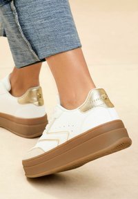 Adidași albi cu un accent auriu la toc, având o talpă groasă de culoare maro, material texturat și un design perforat pe laterale.