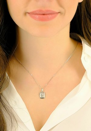 Collier en argent avec un pendentif rectangulaire présentant un motif gravé. La chaîne est fine et délicate, portée sur une chemise blanche à col.