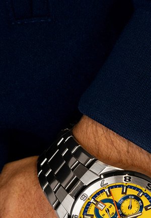 Montre en acier inoxydable argenté avec un cadran jaune et bleu, présentant de grands index horaires et un bracelet à maillons, portée au poignet.