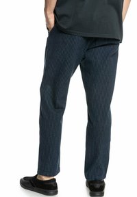 Marinblå randiga byxor i ett tyg med en slät yta, som har en avslappnad passform. Bärs med svarta slip-on skor.