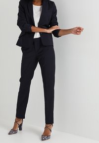 Námořnický blazer a kalhoty na míru, doplněné bílým topem. Úzký střih, hladká textura. Lodičky na vysokém podpatku s květinovým vzorem a kotníkovými pásky.