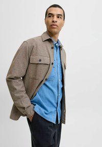 Brun tekstureret jakke med knap-lukkede brystlommer, lagdelt over en lyseblå denimskjorte, parret med mørke bukser. Neutral baggrund.