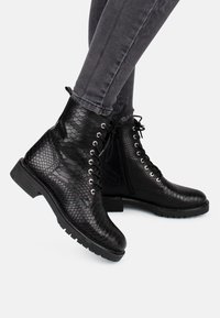 Bottes de cheville noires avec une texture en cuir de serpent, dotées d'œillets argentés, de lacets noirs et d'une semelle en caoutchouc robuste. Finition avec des coutures détaillées.