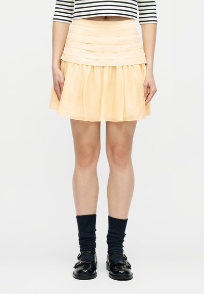 Γυναίκα που φορά κοντή πλισέ φούστα σε απαλό κίτρινο, μαύρα μοκασίνια με αγκράφες, μπλε κάλτσες μέχρι τη μέση της γάμπας και μαύρο-λευκό ριγωτό κοντό τοπ.