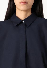 Camisa de color azul marino con cuello y cierre de botones, que presenta una textura suave y líneas limpias sin patrones ni acentos.
